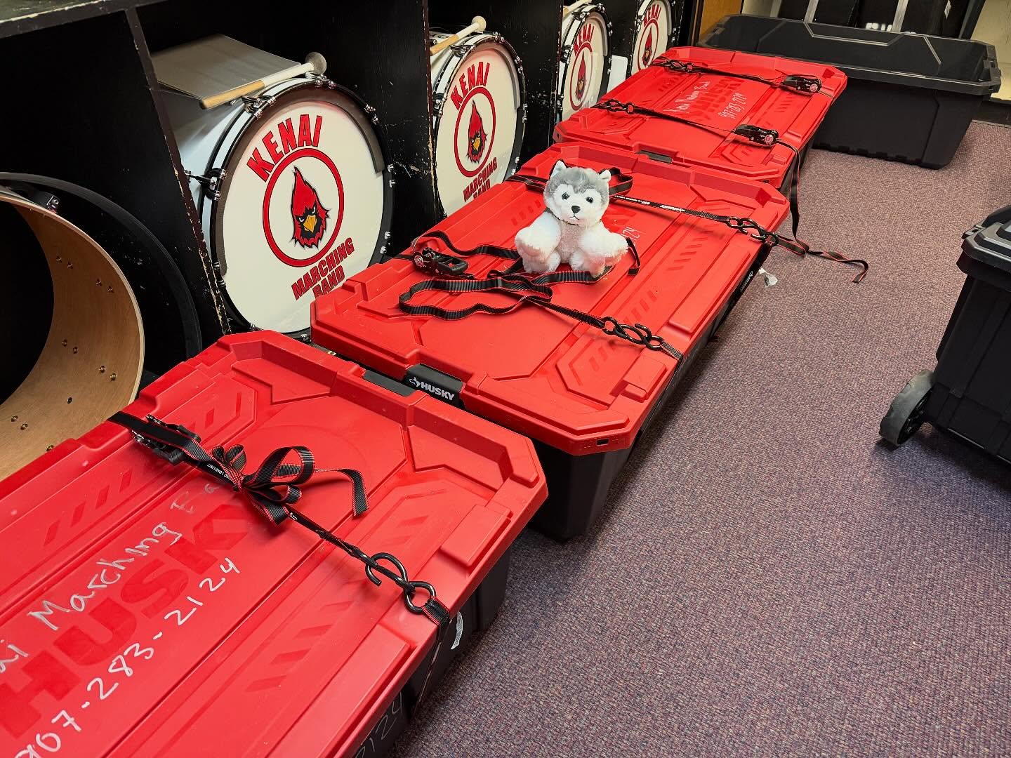 A row of plastic storage totes with red lids. A husky stuffed animal sits on one of the totes.
