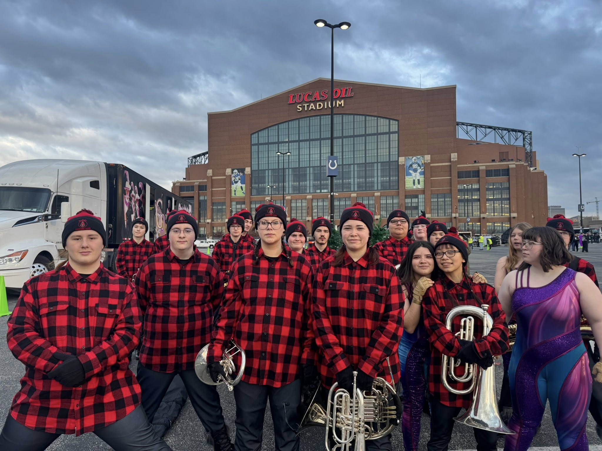 Band students wearing black and red checked flannels and holding instruments pose for a photo in front of Lucas Oil Stadium
