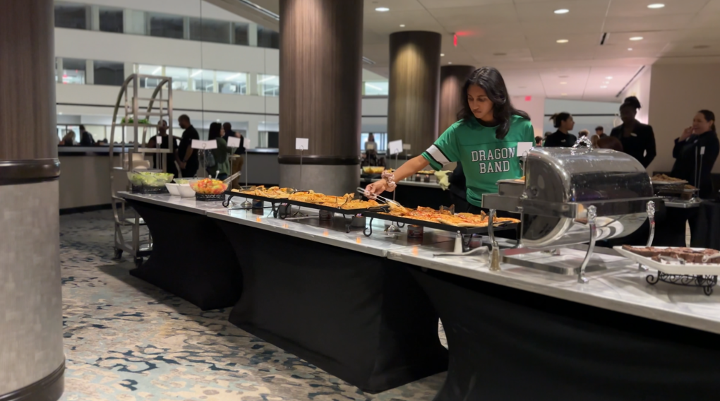 Student in a green shirt takes a slice of pizza from a long buffet table holding salad, fruit salad, several trays of pizza and brownies.