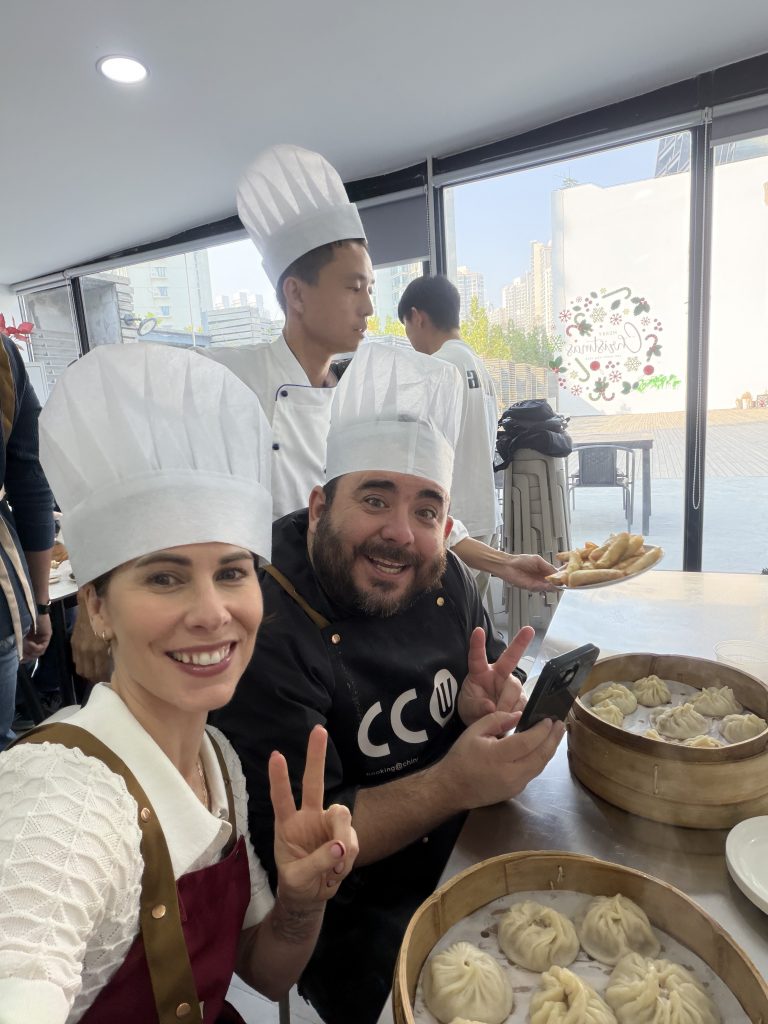 Mary Godley and a man wear chef hats and pose during a dumpling-making class